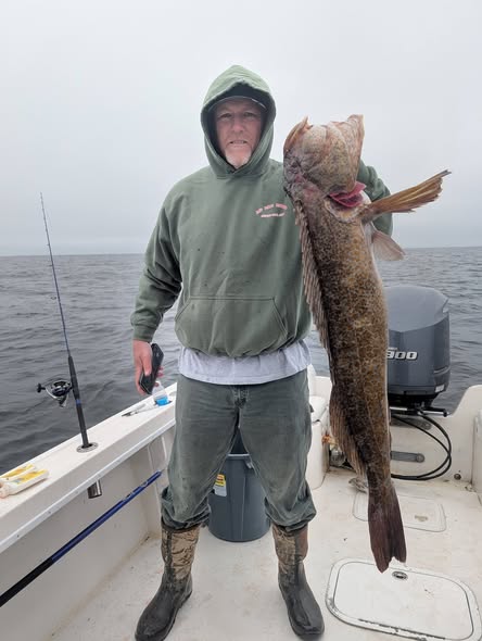 A fisherman in a green hoodie holds up a large catch, showcasing a hefty fish against a grey, overcast ocean backdrop.