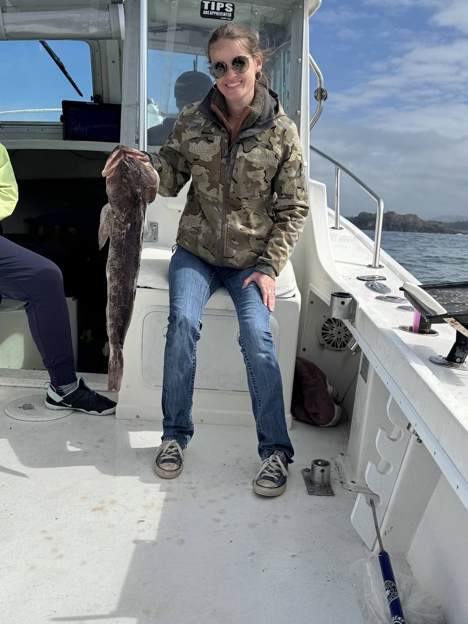 Woman in camouflage jacket smiles while holding a large fish on a boat, showcasing a successful fishing trip in a coastal setting.