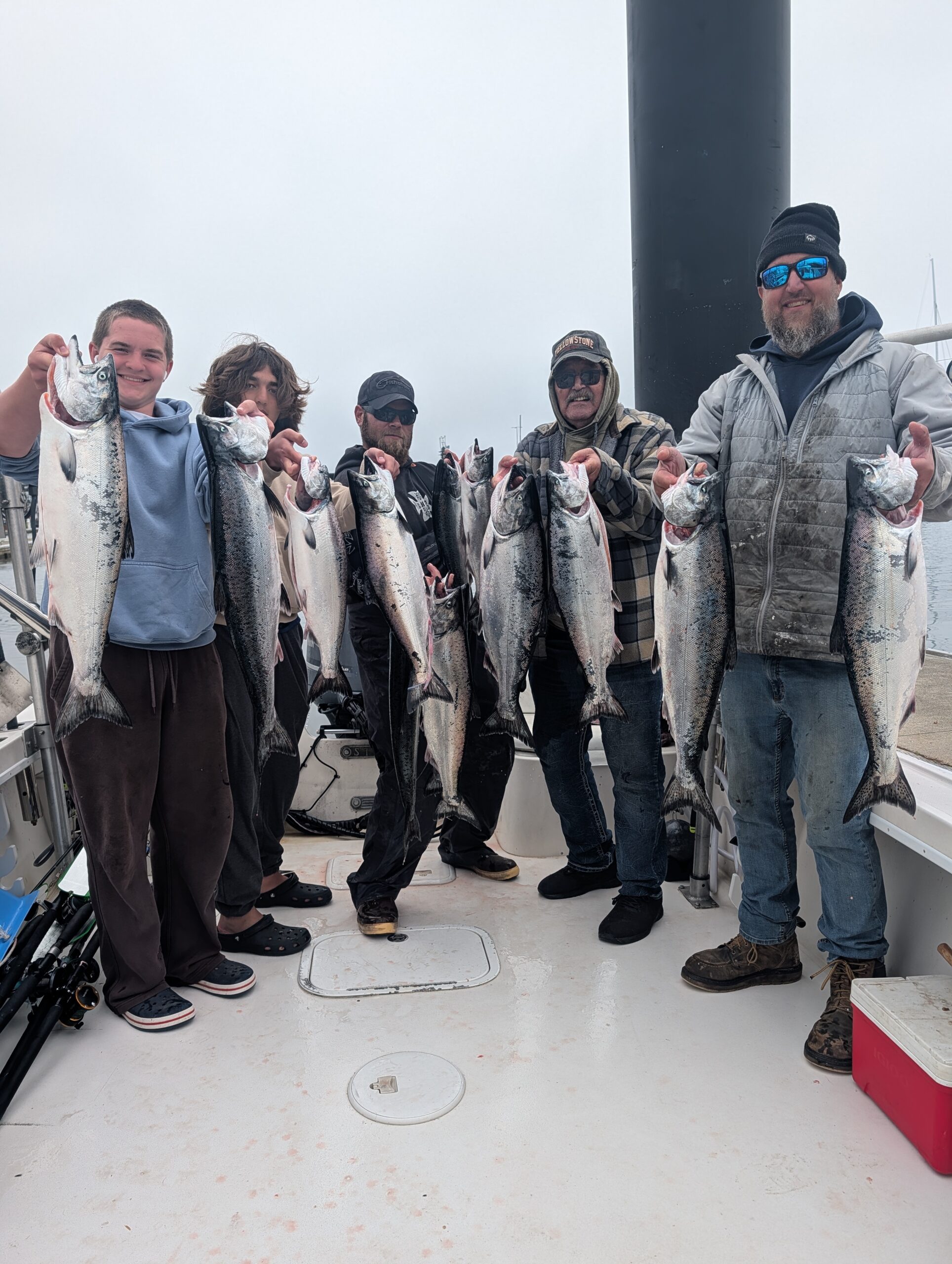Five fishermen proudly display their catch of salmon on a boat, showcasing their successful fishing trip in a foggy coastal setting.