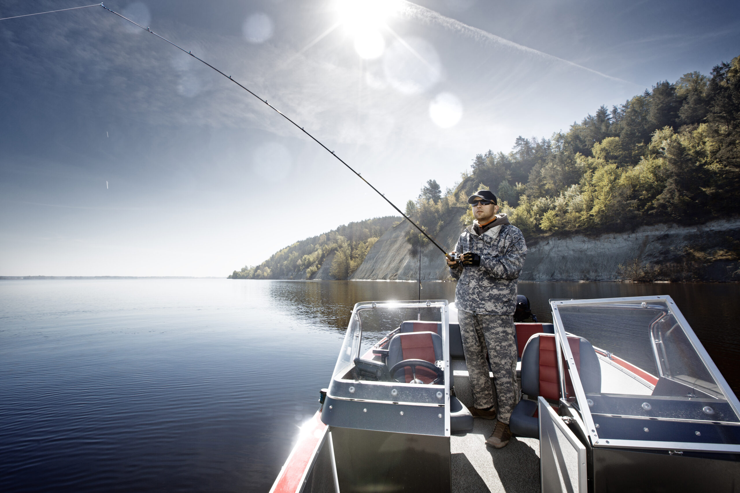 A fisherman in camo attire stands on a boat, casting his line into a serene lake under a bright sky, surrounded by lush green hills.