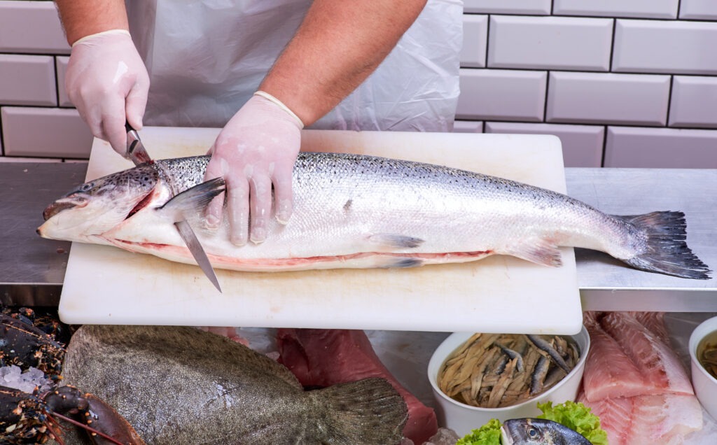 A person in gloves skillfully fillets a large fish on a cutting board, surrounded by various seafood. This image illustrates fish preparation techniques in a culinary context.